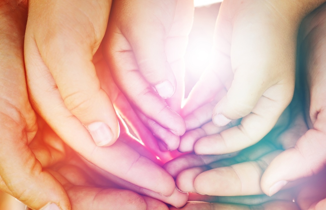 Several adult and child hands are gently overlapping and cupping together with sunlight shining through the center.