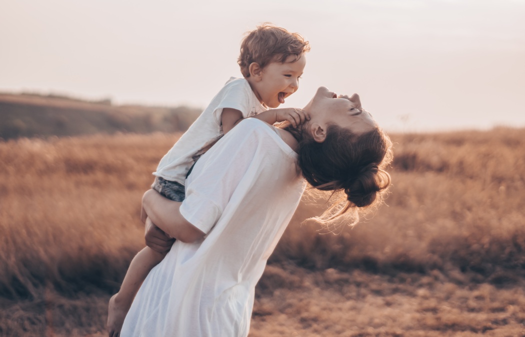A woman in a white shirt holds a young child outdoors in a field at sunset; both appear joyful and are facing each other.