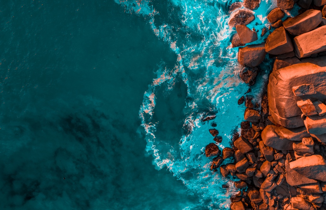 Aerial view of turquoise ocean waves crashing against a rocky, orange-toned coastline.