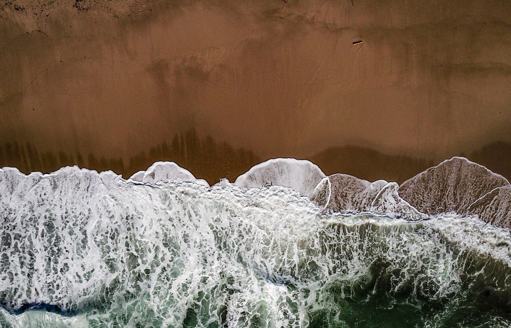 Aerial view of ocean waves crashing onto a sandy beach, creating white foam along the shoreline.