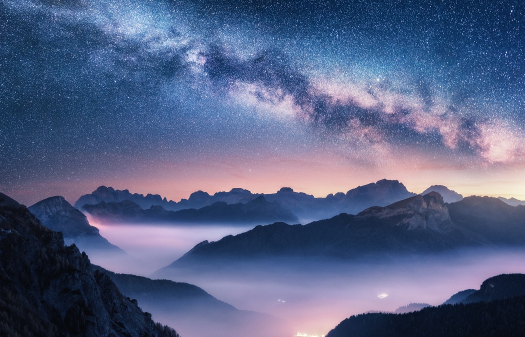 A clear night sky with the Milky Way stretching above mountain silhouettes and a valley filled with misty fog, illuminated by faint lights below.
