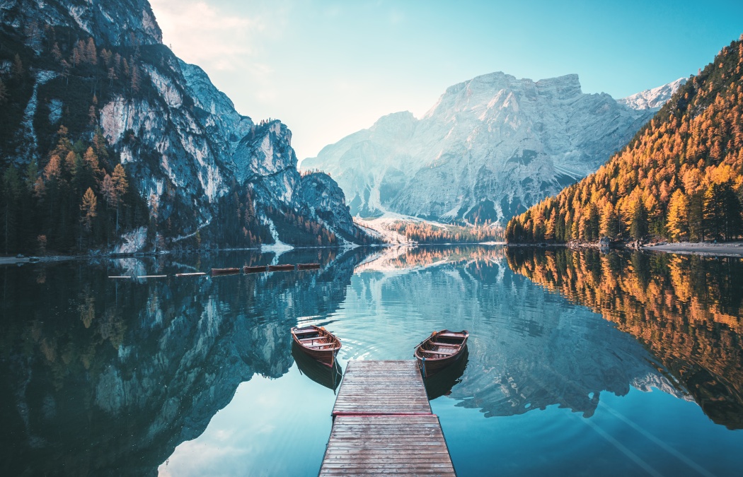 A wooden dock with two rowboats extends into a calm mountain lake, surrounded by forested slopes and high peaks reflected in the water.