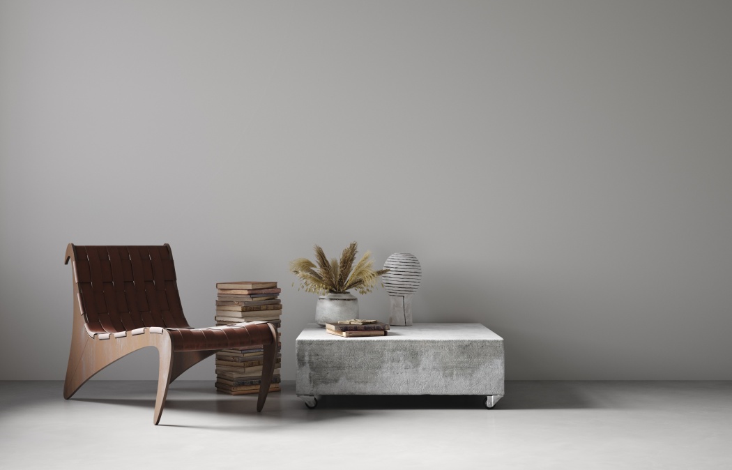A modern minimalist room with a brown woven chair, stacked books, a concrete coffee table, a potted plant, and a striped decorative object against a plain light gray wall.