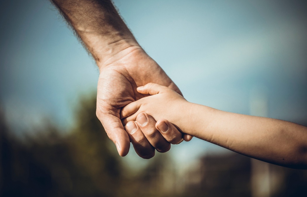 An adult hand holding a child's hand outdoors, with a blurred background.