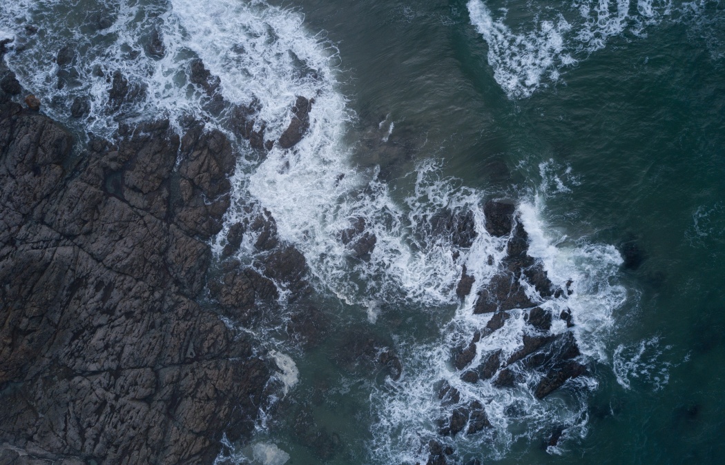 Aerial view of waves crashing against dark, jagged rocks along a coastline, with white foam and deep green water.