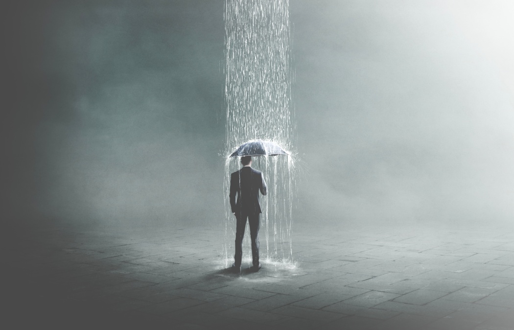 A person in a suit stands on a stone surface under an umbrella, with rain falling in a narrow column directly above them.