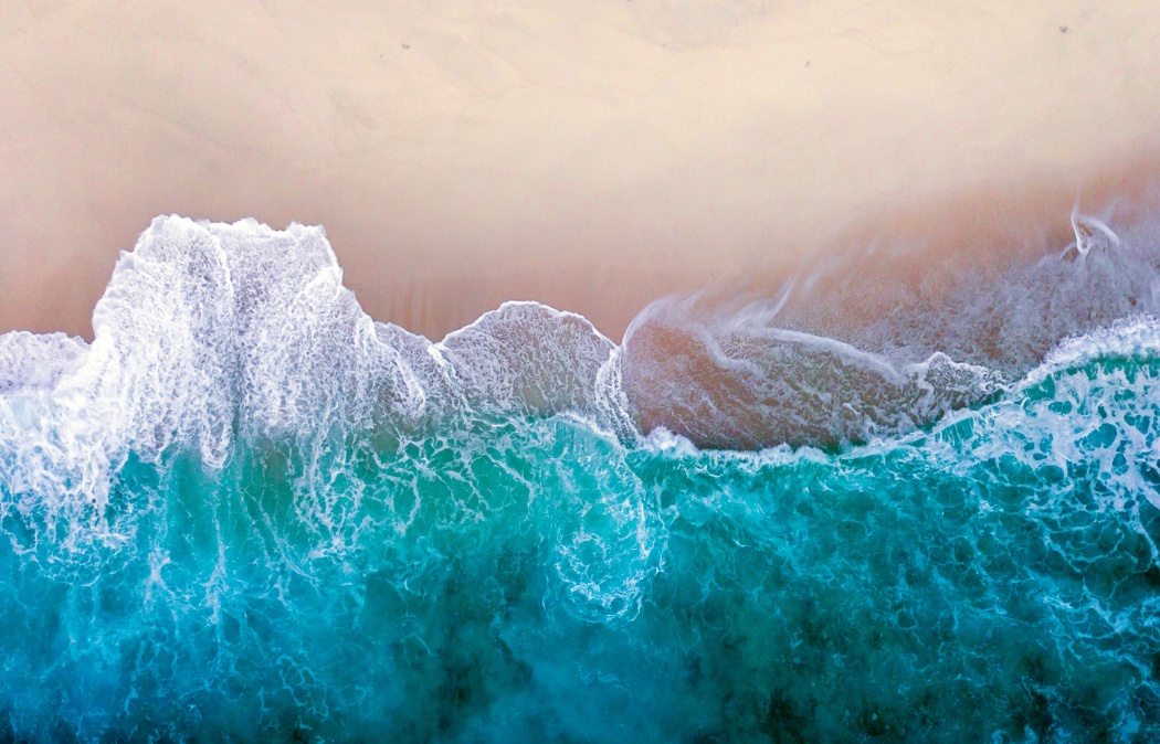 Aerial view of ocean waves crashing onto a sandy beach, with white foam contrasting against blue-green water and smooth beige sand.