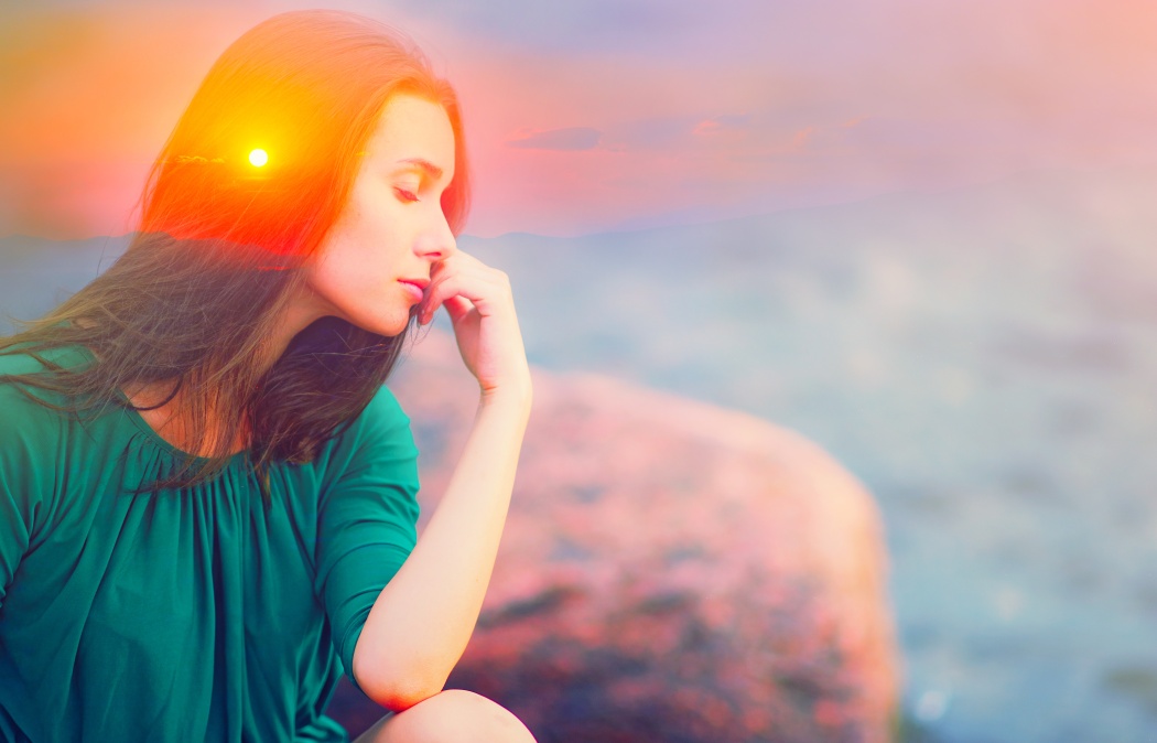 A woman in a green dress sits with her eyes closed and hand near her mouth. Sunlight and colorful haze overlay the scene, with rocks and water in the background.