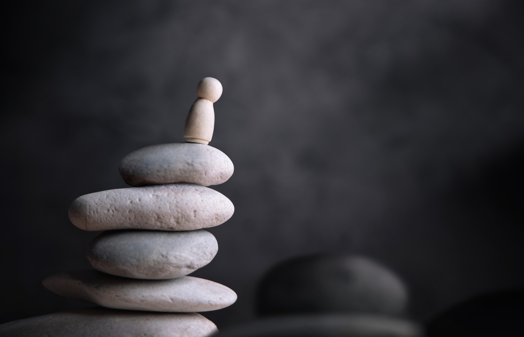 A small wooden figurine stands on top of a stack of smooth, rounded stones against a dark, blurred background.