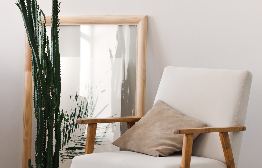 A tall cactus stands next to a framed botanical print and a beige armchair with a tan cushion in a minimalist room.