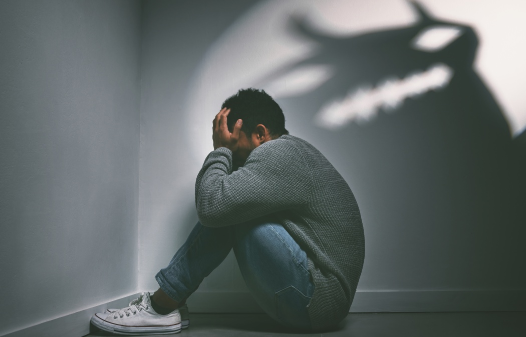 A person sits on the floor in a corner with their head in their hands, while a shadow resembling a monster is cast on the wall behind them.