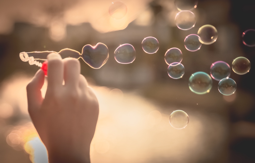 A hand holds a bubble wand shaped like a heart, blowing several soap bubbles against a softly blurred background.