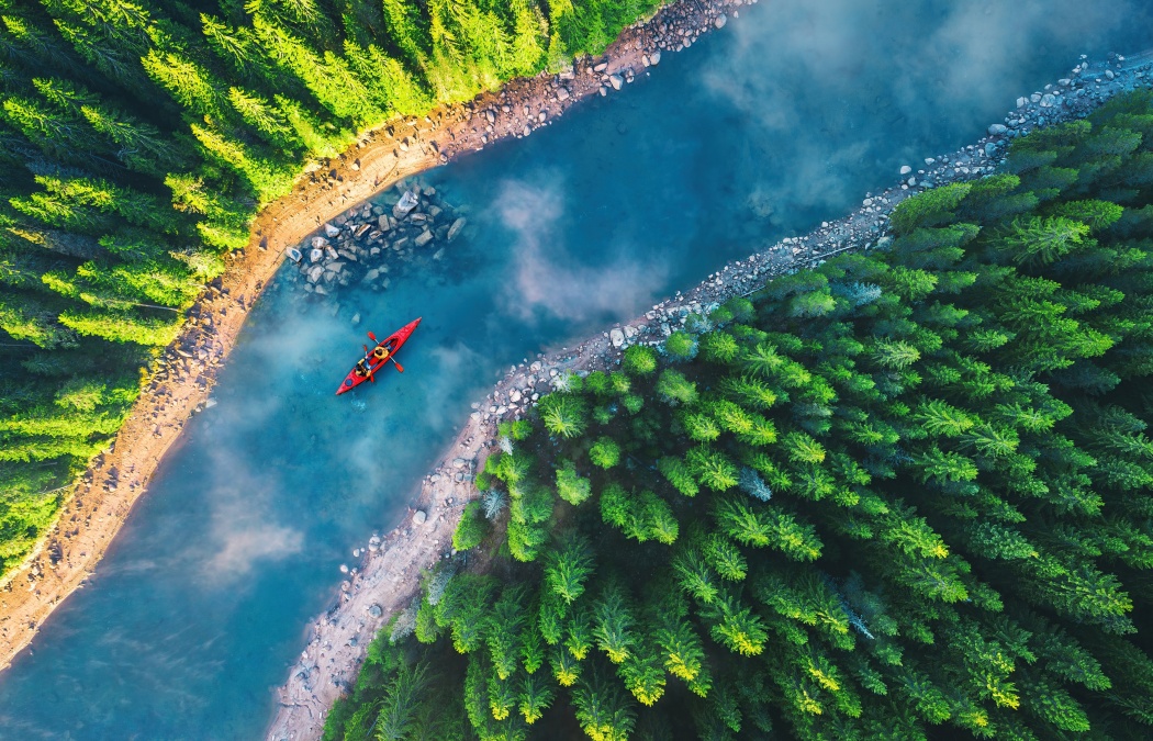 A red kayak with two people paddles on a winding blue river surrounded by dense green pine trees, viewed from above.