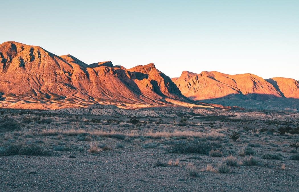 Desert landscape with dry, rocky ground in the foreground and sunlit mountains in the background under a clear sky.