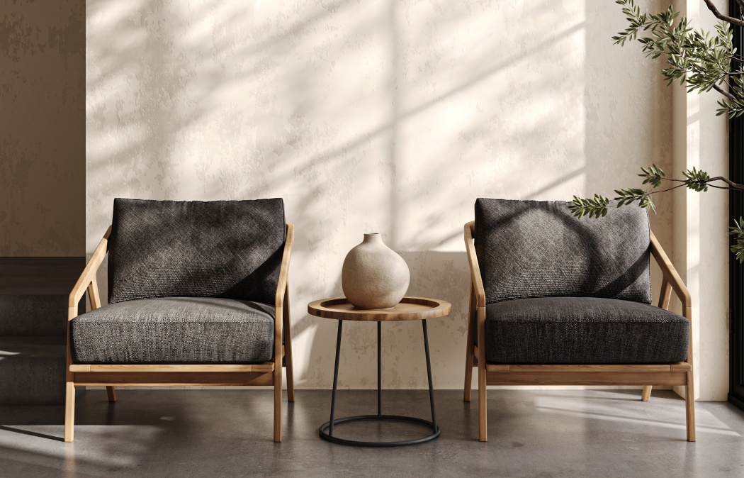 Two modern wooden armchairs with dark cushions are positioned on either side of a small round table with a ceramic vase, against a light textured wall with sunlight streaming in.