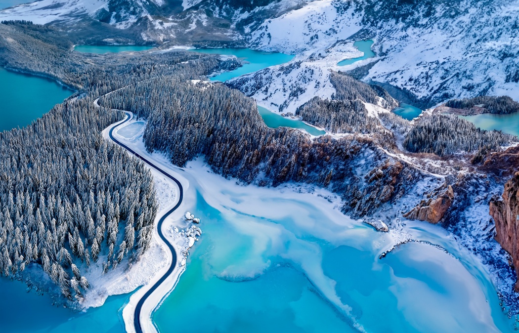 A winding road curves through a snow-covered forest beside turquoise lakes, with mountains in the background under a clear sky.