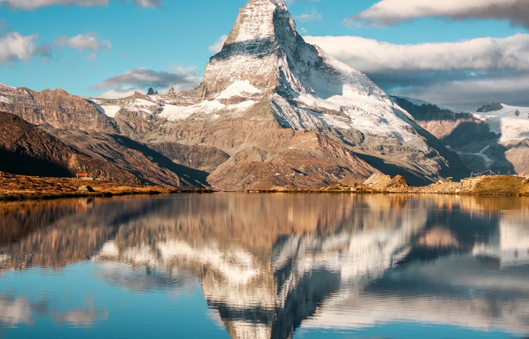 Snow-capped mountain reflected in a calm lake under a partly cloudy sky, with rocky terrain in the foreground.