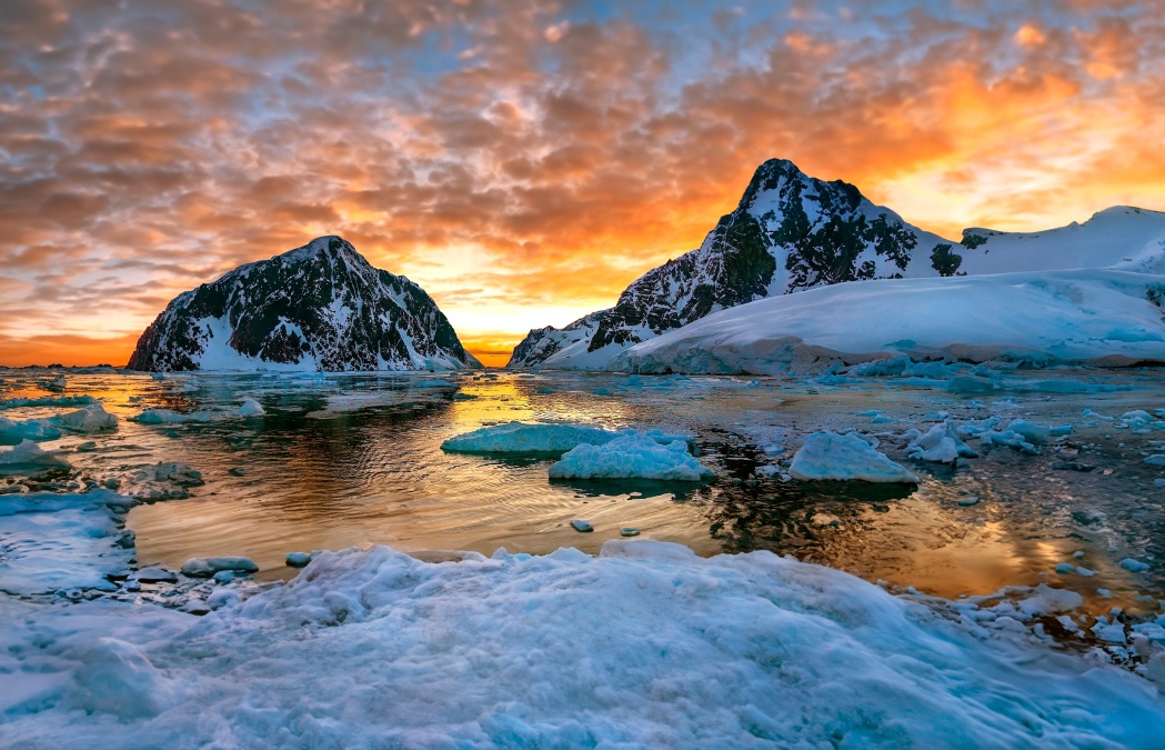 Snowy mountains and icebergs reflected in a calm body of water at sunset, with dramatic orange and yellow clouds filling the sky.