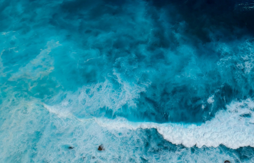 Aerial view of ocean waves crashing, showing varying shades of blue water and white sea foam.