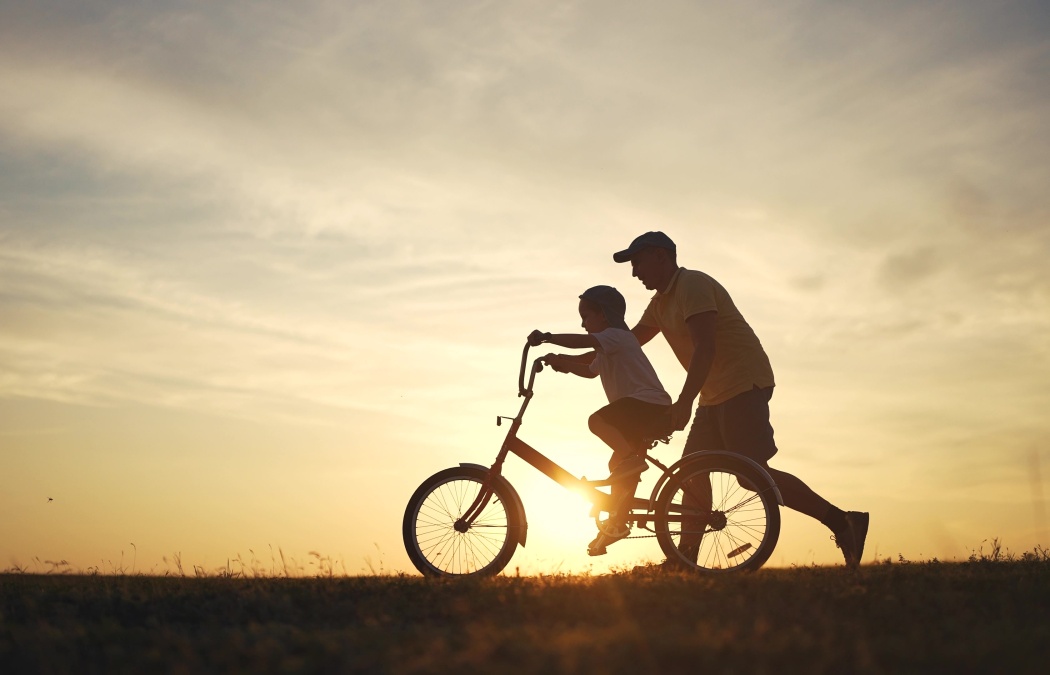 Silhouette of an adult helping a child ride a bicycle at sunset in an open field.