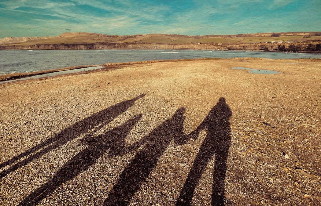 Four long shadows of people standing on a rocky beach, facing towards the water and distant hills under a blue sky.