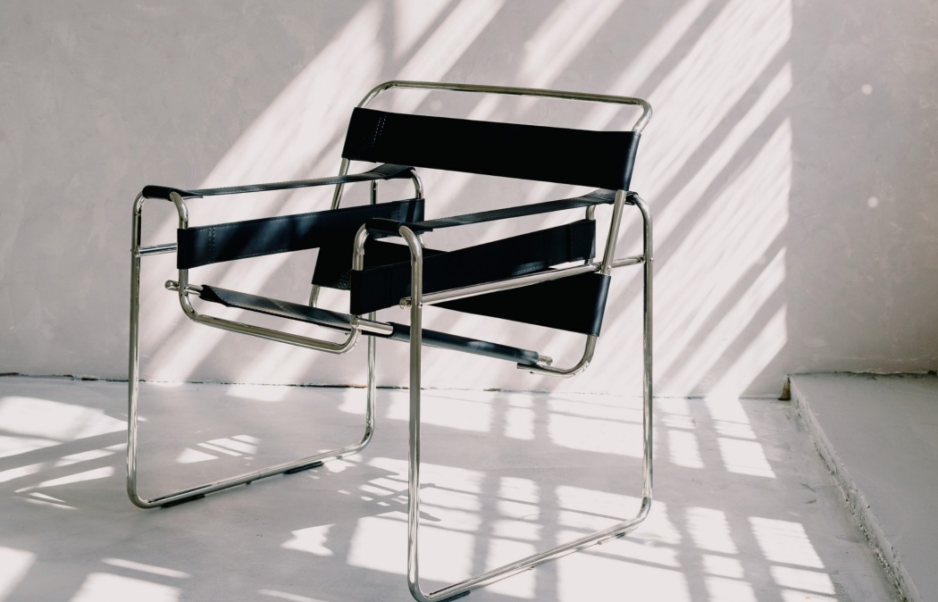 A modern chair with a chrome frame and black seat and backrest sits on a sunlit white floor, with striped shadows cast across the scene.