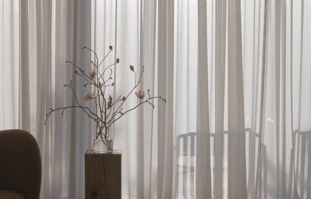 A small glass vase with delicate branches and flowers sits on a wooden table in front of sheer white curtains, next to a brown chair.