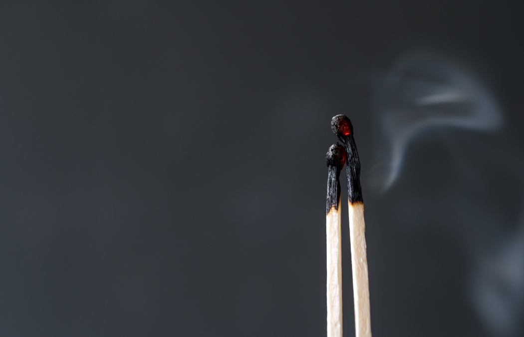 Two burnt matchsticks with blackened heads lean against each other, with faint smoke rising against a plain dark background.