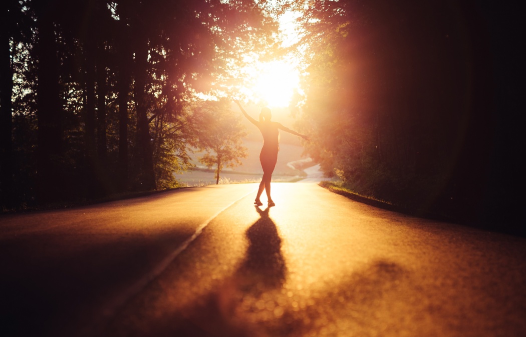 Silhouette of a person standing on a sunlit road with arms outstretched, surrounded by trees, during sunrise or sunset.