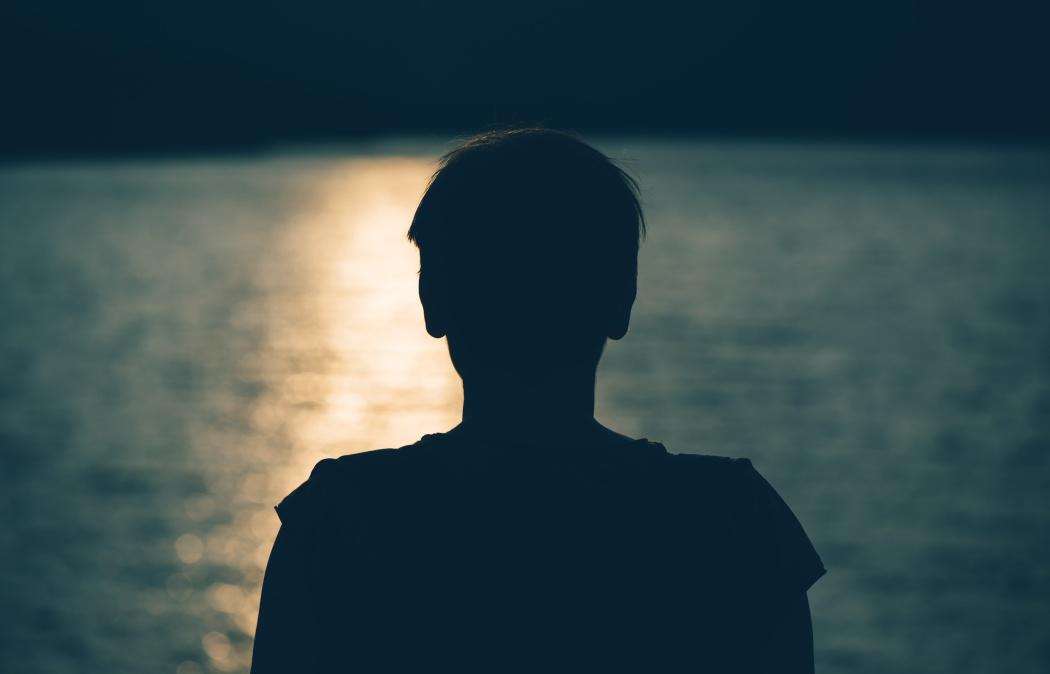 Silhouette of a person facing a body of water at sunset, with sunlight reflecting off the water’s surface.