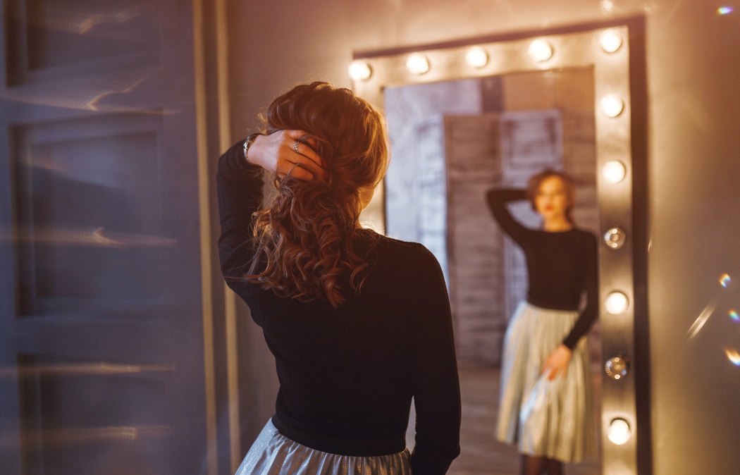 A woman with wavy hair, wearing a black top and metallic skirt, stands in front of a large, lit mirror, holding her hair with one hand and looking at her reflection.