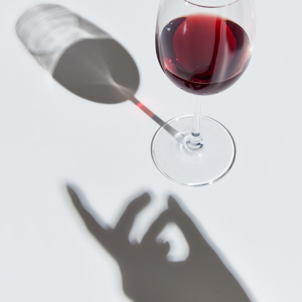 Glass filled with delicious red wine, standing against white background with female hand shadow. Celebration. Concept of taste, alcohol, wine degustation, variety, winemaking. Flat lay