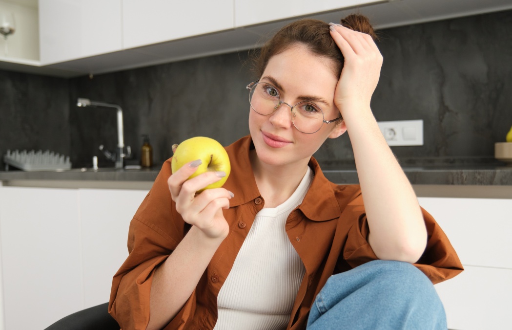 Portrait of smiling, modern woman in glasses, sitting at home and eating green apple, having a lunch break, biting fruit.