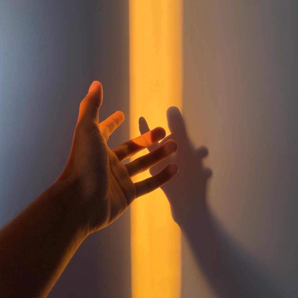 A hand reaching out towards a warm beam of light with a shadow cast on the wall, creating a dramatic and introspective scene. Ideal for projects focusing on light, shadows, and human connection