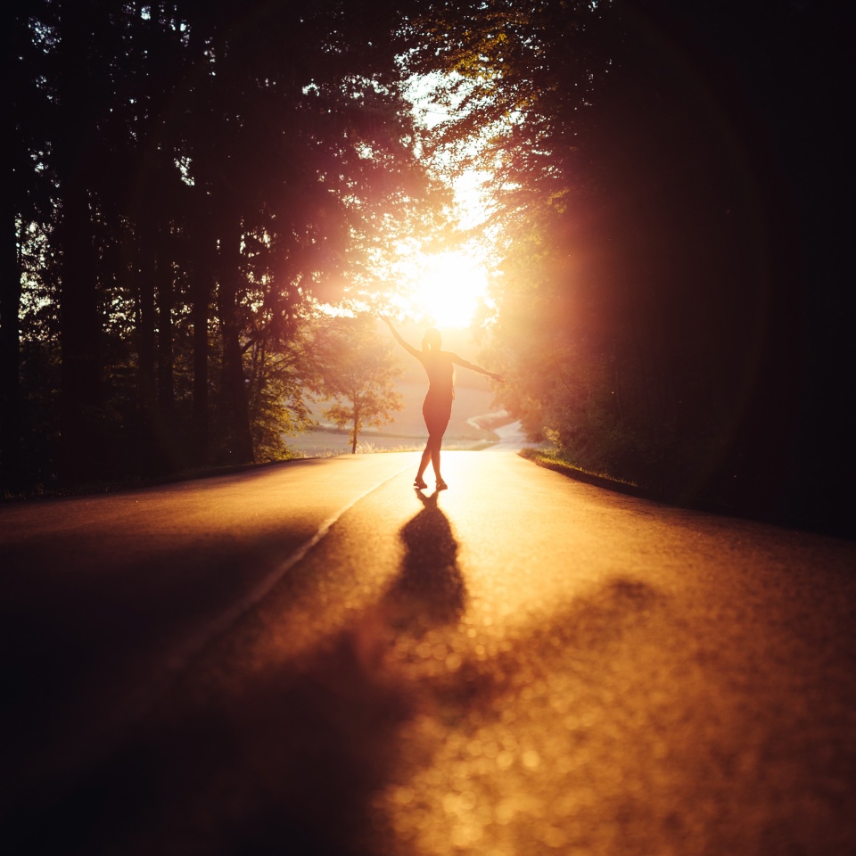 Silhouette of woman captured in relaxed pose with arms outstretched and crossed legs. In the background is the glow of the sun above the horizon, illuminating woman and casting her shadow on the road.