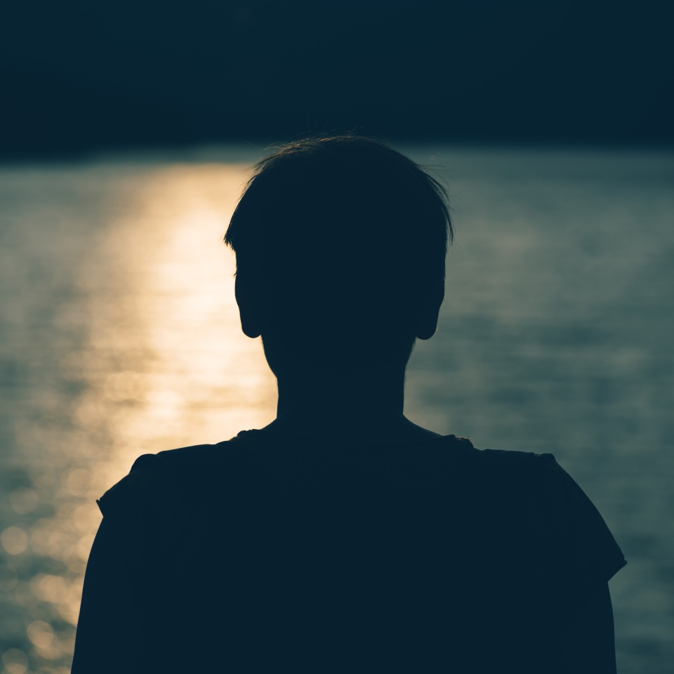 Silhouette of depressed sad woman standing by the lake in cold autumn sunset. Gloomy adult female person looking at water. Loneliness and solitude concept.