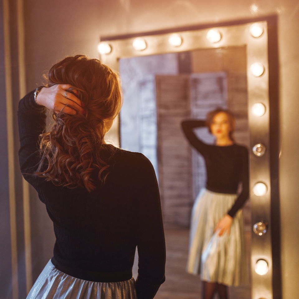 Woman in a beauty salon looks at her reflection in the mirror with lamps and checks hairstyle and makeup