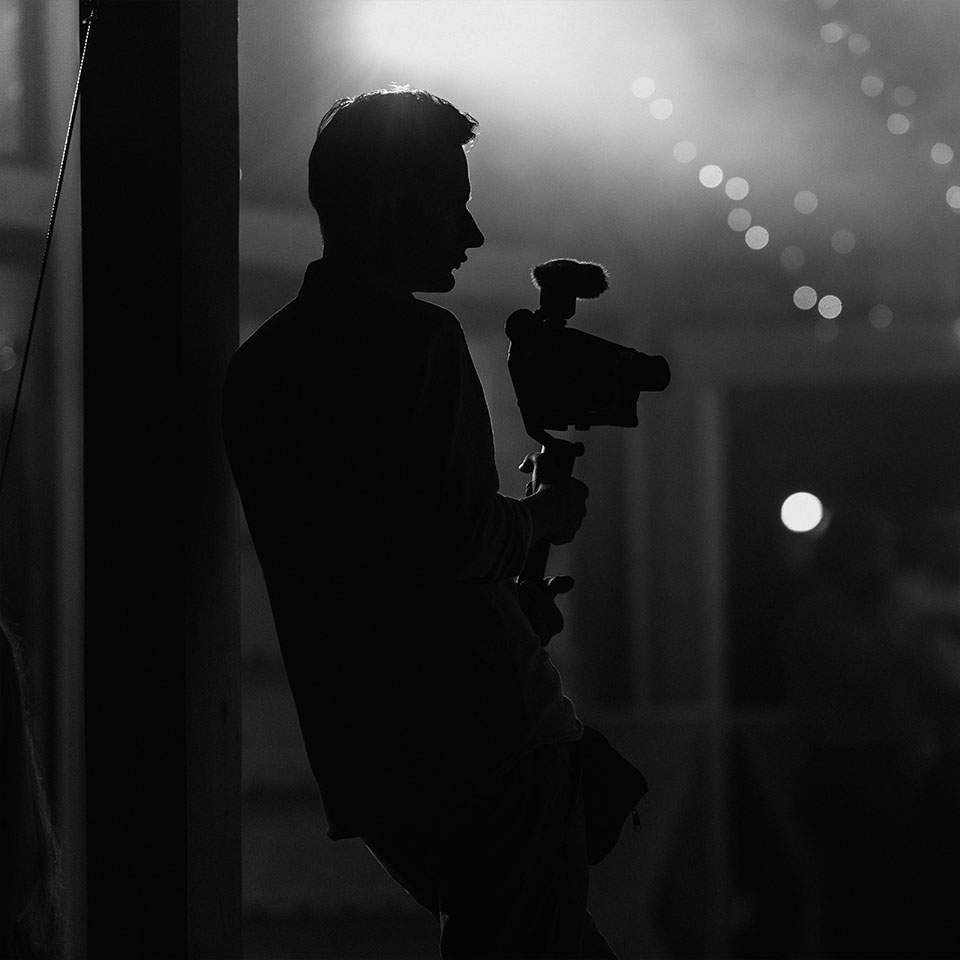 Silhouette of a person leaning against a post while holding a camera with a microphone; background is dimly lit with some blurred lights.