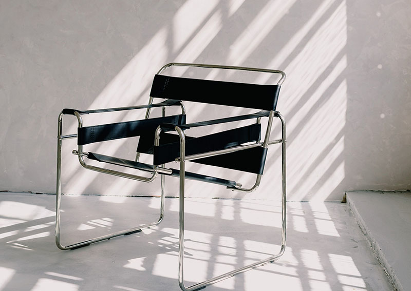 A modern chair with a metal frame and black leather straps sits on a white floor, with shadows from window blinds cast on the wall behind it.