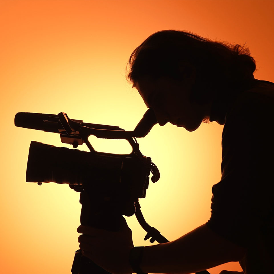 Silhouette of a person operating a professional video camera against an orange-yellow backlight.
