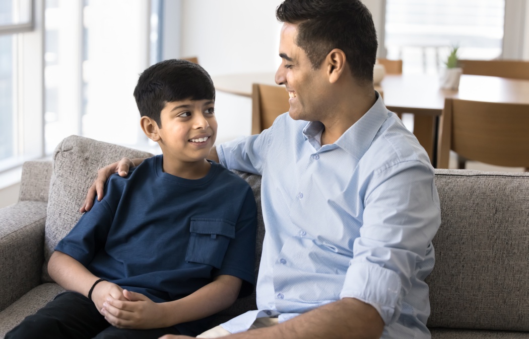 Happy Indian preteen son talking to loving father on home sofa. Positive caring dad enjoying friendship, good relationship with teenage kid, giving support to boy, hugging child