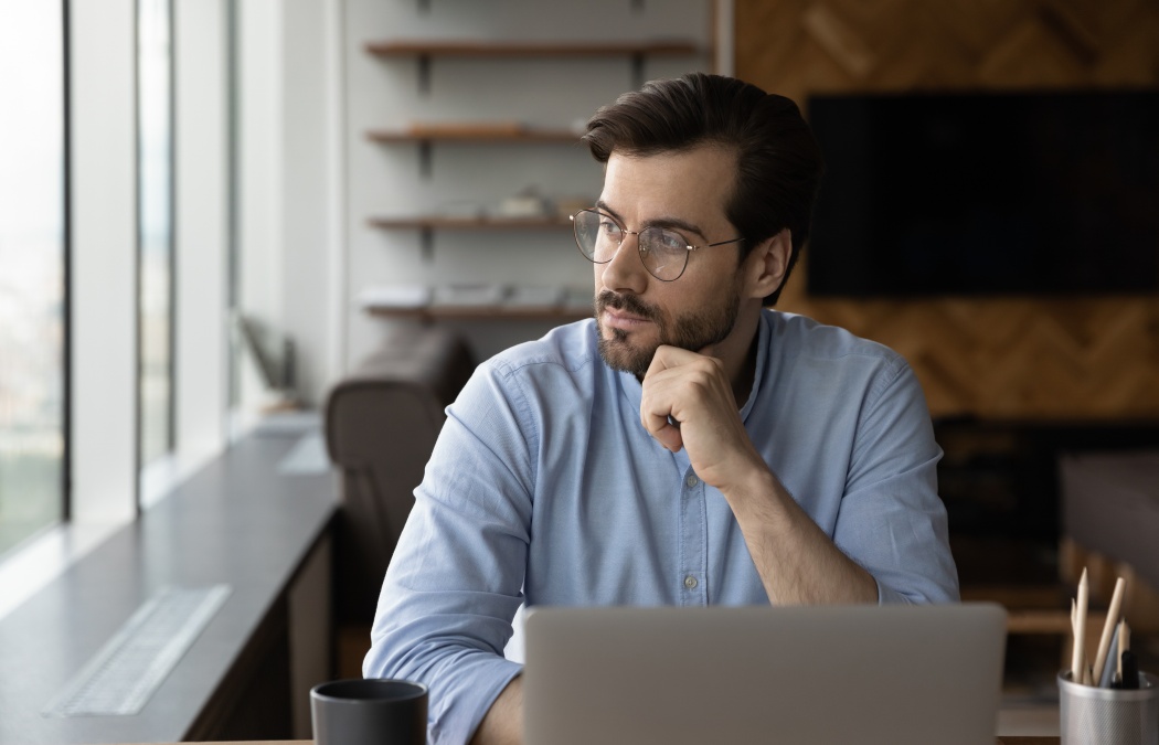 Thoughtful businessman in glasses looking at window from workplace with laptop, thinking over challenges, work issues, problem solving, making decision, feeling doubts, pondering on future vision